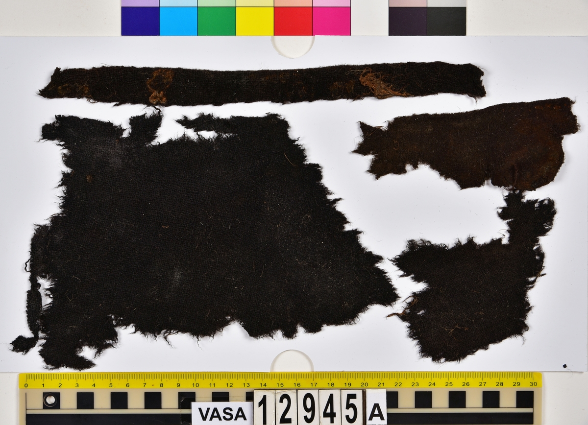 Textilfragment.
15 textilfragment uppdelade på fyndnummer 12945a-g.
Fnr 12945a består av fyra fragment av ull vävda i tuskaft. Två av fragmenten har rester av söm eller fåll samt har varit valkade.
Fnr 12945b består av två fragment av ull vävda i tuskaft. 
Fnr 12945c består av ett fragment av ull vävt i tuskaft. Fragmentet kan ha varit valkat på ena sidan.
Fnr 12945d består av ett fragment av ull vävt i tuskaft. Fragmentet har en söm med rester av stygn kvar i ena kanten.
Fnr 12945e består av tre fragment av ull vävda i tuskaft. Fragmentet har en tydlig originalkant bevarad.
Fnr 12945f består av ett filtat fragment. Kan möjligen ha varit en del av ett hattbrätte.
Fnr 12945g består av tre filtade fragment, troligen av ull.