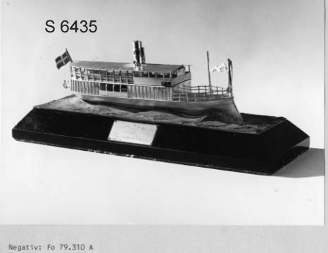 Fartygsmodell av  S/S VICTORIA (1882 vid Bergsund) i silver. Skrov med rammstäv, inbyggd brygga, skorsten med Waxholmsbolagets märke, ingraverat fartygsnamn på stäven, namnvimpel av silver, flagga akter. Livbåt akter. Monterad på vågimiterad kopparplatta och träsockel.