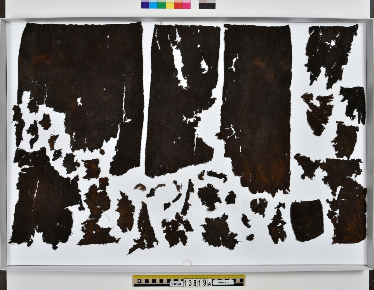 Textilfragment, byxa.
125 fragment uppdelade på fyndnummer 13819a-d.
Fnr 13819a består av 44 fragment av ull vävt i tuskaft och valkat på ena sidan. En del av fragmenten har originalkanter med sömmar och fållar.
Fnr 13819b består av 35 fragment av ull vävt i tuskaft. En del av fragmenten har originalkanter med sömmar.
Fnr 13819c består av 40 fragment av ull vävt i tuskaft. En del av fragmenten har originalkanter med sömmar. Fragmenten är mycket sköra.
Fnr 13819d består av 6 fragment mönstervävt band av ull.