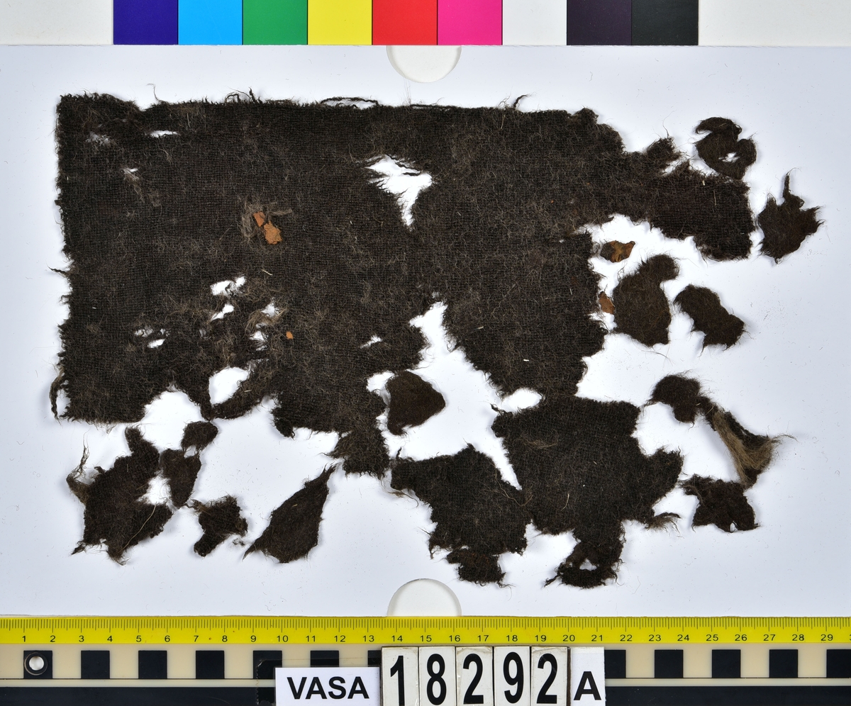 Textil.
26 textilfragment uppdelade på fnr 18292a-b.
Fnr 18292a består av 20 fragment av ull vävda i tuskaft. Ytan har ljusa ullfragment på sig. 
Fnr 18292b består av 6 lösa, ljusa fragment av ull. Samma typ av fragment som på ytan på tyget i 18292a. Ytan kan möjligen ha varit ruggad.