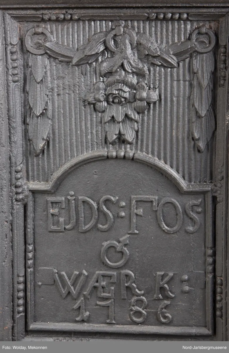 1.etasje kortside, frem:  riflet plate med blomstergirlander, døråpning med buet overkant. Askebrett med trifol og kant rundt. 1.et-kortside, bak: samme som forside, men døråpningen har innskripsjon eidsfos werk 1786 1. et.begge langsider: Riflet plate med prlestaffkant, blomstergirlander over og på sidene med portrett av kvinne i profil i oval medaljong sentrert på platen 2.et. Kortside, frem:  riflet plate med perlestafafkant, blomstergirlander opphengt under feieluke. Blomsterornament over luken. Luken er hamret ut, med låsslå, dekorert med blomsterhode. bak: samme som forsiden, men feieluken markert som rektangulært felt. 2.et.langside buet åpningh kun på langsidene, åpningen har girlander som henger nedenfor åpningen, på siden av åpningen er det bladpilstre. Girlander henger over medaljong med kvinnebyste i profil.