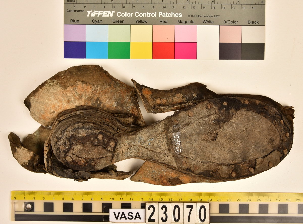 Sko.
Inuti skorna hittades även fragment från olika textilier som ursprungligen varit ett par strumpor. Idag sitter ett stickat fragment av ull fast i vänsterskon (23070) med avigsidan uppåt. I fyndet finns även en mängd lösa stickade fragment som idag är mycket korroderade och vissa helt mineraliserade av järn vilket har gjort textilen stenhård och därmed oerhört skör (23070a-c). Utifrån minskningar som några små fragment har så har det varit möjligt att rekonstruera strumpans tå. Genom att pussla ihop fragmenten som ursprungligen har suttit ihop har det på så vis gått att avgöra hur strumpans tå har varit konstruerad. Då textilen är så fragmentarisk är det inte möjligt att avgöra hur resten av strumpan har sett ut eller varit konstruerad. Däremot så finns det fragment av ett kypertvävt tyg av ull som på vissa ställen ligger dubbelt med det stickade 23070c). Ett av dessa fragment har stelnat i formen så som den suttit på fotens främre del inuti skon. Detta fragment täcker delvis det stickade tyget och har dessutom spår av kaststygn längs en kant. Så som det vävda tyget är placerat på det stickade tyget pekar på att man använt det som lagningslappar till sina stickade strumpor. 
I fyndet finns det även korroderade järnklumpar, ett tuskaftsvävt tyg av ull och rester av ett grovt bastfibergarn och två mindre läderfragment (23070d-f). Det är möjligt att även dessa har tillhört strumpan och/eller skon.

För mer detaljerad information från textildokumentationen (år 2022) se länkade filer.

Skodokumentation 2022:
Ovanläder. Tillverkad i getskinn av god kvalitet. Hör ihop med fyndnummer 23069 (Yngves skor). Tillverkades på en rak läst och bars på vänster fot. Inga dekorationer Det finns stygnhål som visar att det funnits invändiga förstärkningar (men förstärkningslappen saknas). Det finns delaminering men inga lagningar.
För mer information, se Länkade filer: Uppritning

Text in english:
This is the upper of a shoe of more “extreme fashion”, made of goatskin leather of good quality, probably made on a straight last and worn of the left foot, with no decorative features. The upper couples with 23069, together composing Yngve’s shoes. There are internal reinforcements missing from the upper and delamination, there are no repairs.
For more information, go to Länkade filer: Uppritning