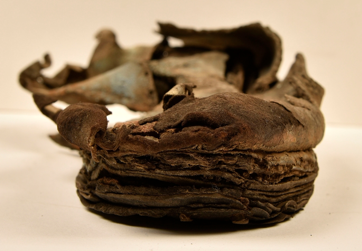 Sko.
Inuti skorna hittades även fragment från olika textilier som ursprungligen varit ett par strumpor. Idag sitter ett stickat fragment av ull fast i vänsterskon (23070) med avigsidan uppåt. I fyndet finns även en mängd lösa stickade fragment som idag är mycket korroderade och vissa helt mineraliserade av järn vilket har gjort textilen stenhård och därmed oerhört skör (23070a-c). Utifrån minskningar som några små fragment har så har det varit möjligt att rekonstruera strumpans tå. Genom att pussla ihop fragmenten som ursprungligen har suttit ihop har det på så vis gått att avgöra hur strumpans tå har varit konstruerad. Då textilen är så fragmentarisk är det inte möjligt att avgöra hur resten av strumpan har sett ut eller varit konstruerad. Däremot så finns det fragment av ett kypertvävt tyg av ull som på vissa ställen ligger dubbelt med det stickade 23070c). Ett av dessa fragment har stelnat i formen så som den suttit på fotens främre del inuti skon. Detta fragment täcker delvis det stickade tyget och har dessutom spår av kaststygn längs en kant. Så som det vävda tyget är placerat på det stickade tyget pekar på att man använt det som lagningslappar till sina stickade strumpor. 
I fyndet finns det även korroderade järnklumpar, ett tuskaftsvävt tyg av ull och rester av ett grovt bastfibergarn och två mindre läderfragment (23070d-f). Det är möjligt att även dessa har tillhört strumpan och/eller skon.

För mer detaljerad information från textildokumentationen (år 2022) se länkade filer.

Skodokumentation 2022:
Ovanläder. Tillverkad i getskinn av god kvalitet. Hör ihop med fyndnummer 23069 (Yngves skor). Tillverkades på en rak läst och bars på vänster fot. Inga dekorationer Det finns stygnhål som visar att det funnits invändiga förstärkningar (men förstärkningslappen saknas). Det finns delaminering men inga lagningar.
För mer information, se Länkade filer: Uppritning

Text in english:
This is the upper of a shoe of more “extreme fashion”, made of goatskin leather of good quality, probably made on a straight last and worn of the left foot, with no decorative features. The upper couples with 23069, together composing Yngve’s shoes. There are internal reinforcements missing from the upper and delamination, there are no repairs.
For more information, go to Länkade filer: Uppritning