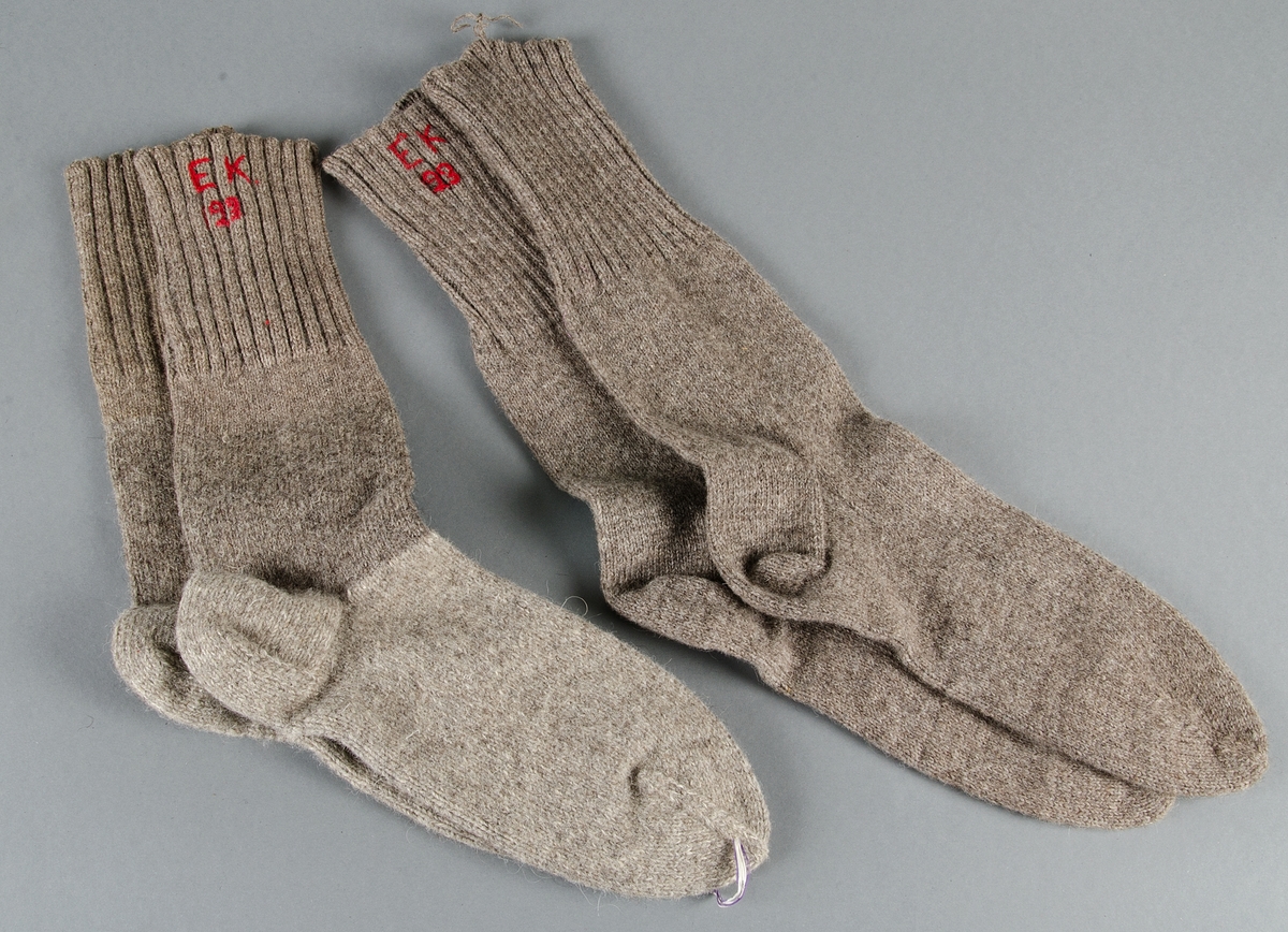 Två par stickade yllesockor. Långa skaft. Det ena paret sitter ihop med lila/vitt paketsnöre. Ett par ljust brungrå och ett med med ljust brungrå skaft och ljusgrå fot. Broderat monogram EK samt beväringsnumret 123 i rött. En socka saknar monogrammet. I dess skaft sitter en ulltråd med vilken paret suttit ihop.