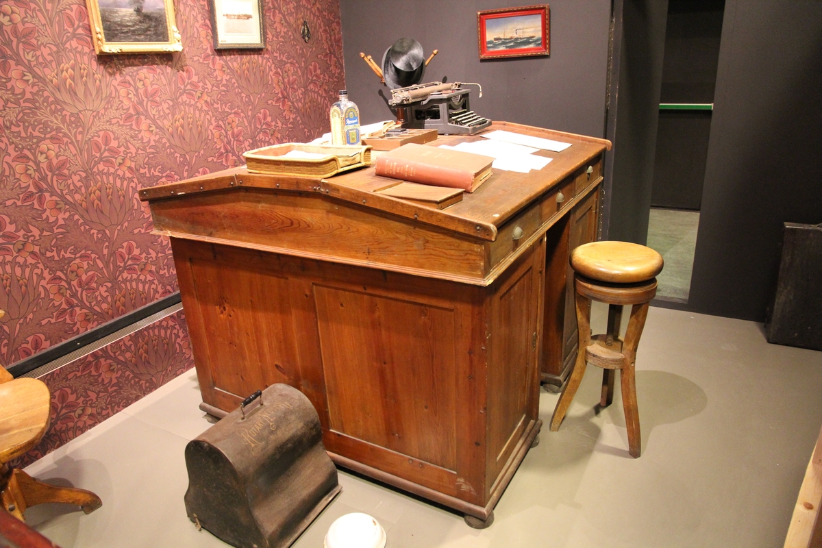 Skrivepult til å stå ved, på hver side to skapdører med åpning i midten. Over dette 3 skuffer . Likt på begge  sider. Den skrå platen med skuffer kan løftes av. Låskasse og håndtak i metall.