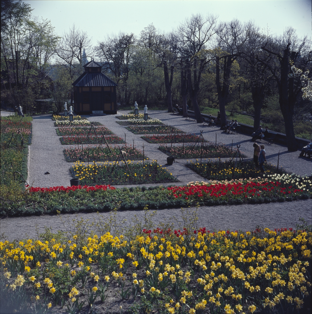 Blommande vårblommor i Rosenträdgårdens välskötta rabatter. I bakgrunden begrundar Swedenborgs lusthus fyra figurer som dansar i den färgrika blomsterprakten.