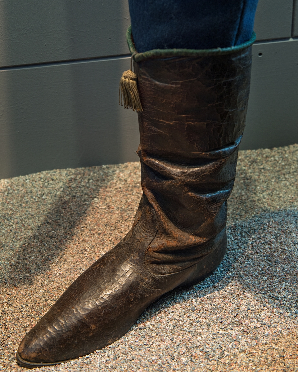 Stövlar, svartlackerade, korta skaft med guldtofs på framsidan, kantade med grönt läder. Sydda, smal fot och spetsig tå. Tillhörande järnskena för fästande till mannekäng.