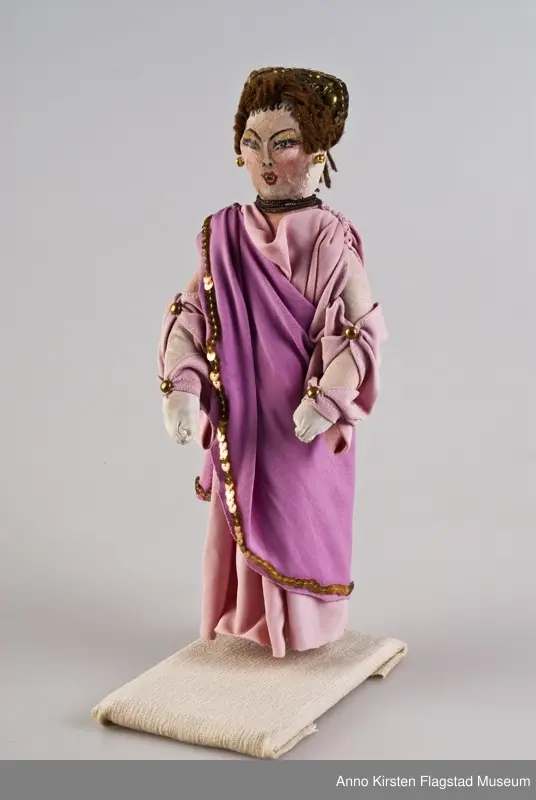 Kvinneskikkelse i operakostyme: Dido fra Henry Purcells opera "Dido og Aenaes". Lys, og mørk syrinfarvet lang kjole med vide ermer. Holdes sammen av fire forgylte knapper. Kjolen har border av forgylte paljetter og forgylt spenne på høyre skulder. Forgylt og grønn hodepynt. Hår av brunt ullgarn. Forgylte perler i ørene og treraders kjede. Malt ansikt i sort, grønt, rosa og rødt. 