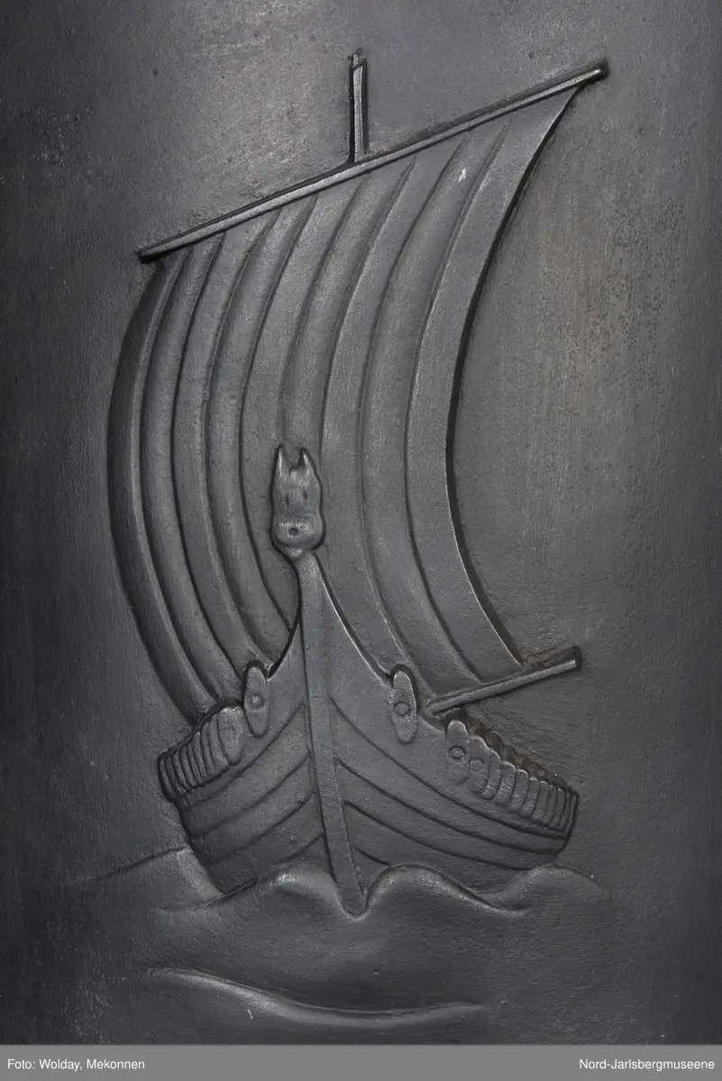 Vikingscene med fem vikinger og to skip, i front. PÅ toppstykket tre øksehoder på rad, vendt inn mot en  midtplassert medaljong med drageornament. Kokerommets kortsider relieff med vikingskip sett rett forfra for fulle seil.