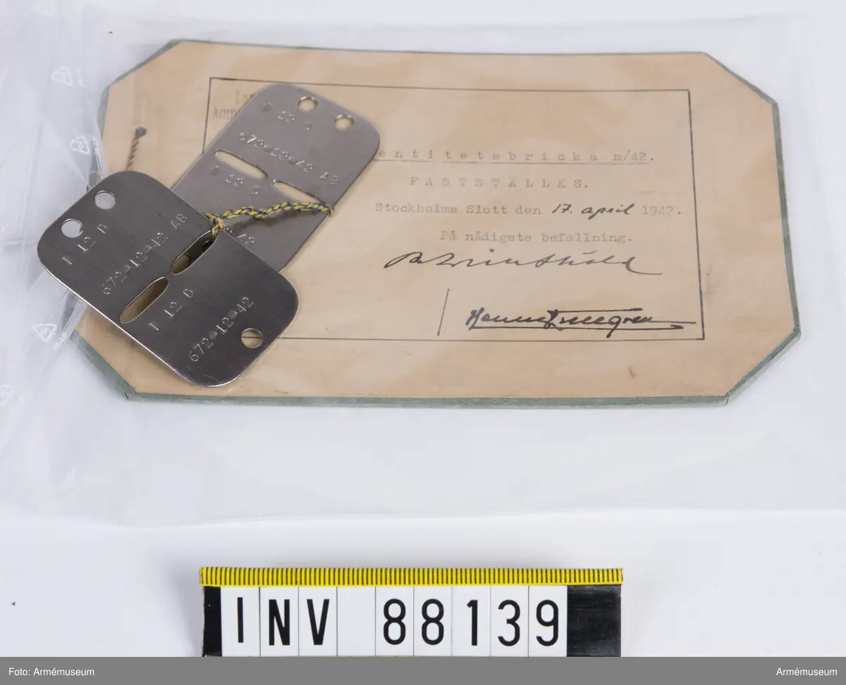 Två stycken identitetsbrickor m/1942 präglade med text: "I 12 D 672-12-42 AB". Vidhängande modellapp med text: "Lantförsvarets kommandoexpedition N:r 1519. Modell å identitetsbricka m/42. Fastställes. Stockholms slott den 17 april 1942. På nådigaste befallning. (oläsliga signaturer)."