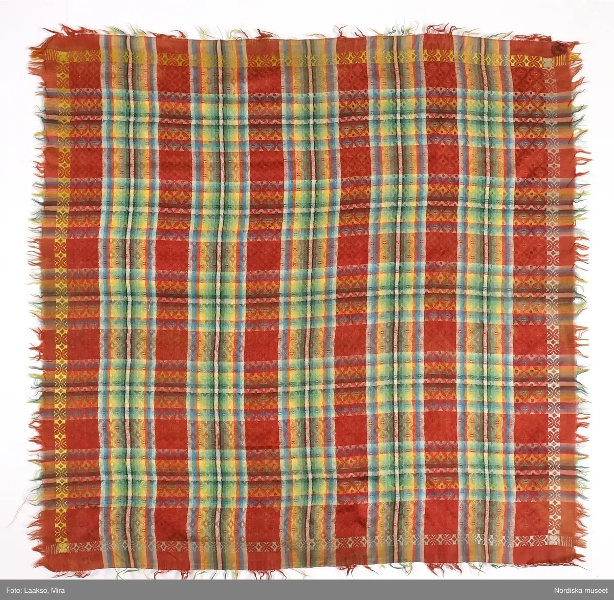 Kvadratiskt halskläde av siden, orangeröd botten med rutning i gult, grönt, ljusblått och vitt samt en smal ytterbård med stiliserad blomma längs 2 sidor i vitt och längs de andra i gult. Botten mönstervävd i ett bindemönstermönster med kryss och små stjärnor, i ytterkant utanför bården slät tuskaft. Uppsrispad kort frans runt alla sidor. I ena hörnet en rund svart hallstämpel med texten. Stockholm 1825 och i mitten St. Eriksbild. Troligen vävd i en skaftvävstol.
Typen var vanlig på 1820-talet och finns från flera håll i landet bl.a 98530 från Äppelbo och 93669 från Malung  i Dalarna. även 44279 från Västmanland och 91632, 152104, 162623 från Småland.
I fint skick
/Berit Eldvik 2011-11-23