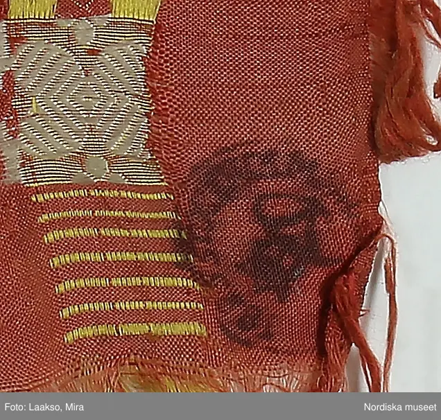 Kvadratiskt halskläde av siden, orangeröd botten med rutning i gult, grönt, ljusblått och vitt samt en smal ytterbård med stiliserad blomma längs 2 sidor i vitt och längs de andra i gult. Botten mönstervävd i ett bindemönstermönster med kryss och små stjärnor, i ytterkant utanför bården slät tuskaft. Uppsrispad kort frans runt alla sidor. I ena hörnet en rund svart hallstämpel med texten. Stockholm 1825 och i mitten St. Eriksbild. Troligen vävd i en skaftvävstol.
Typen var vanlig på 1820-talet och finns från flera håll i landet bl.a 98530 från Äppelbo och 93669 från Malung  i Dalarna. även 44279 från Västmanland och 91632, 152104, 162623 från Småland.
I fint skick
/Berit Eldvik 2011-11-23