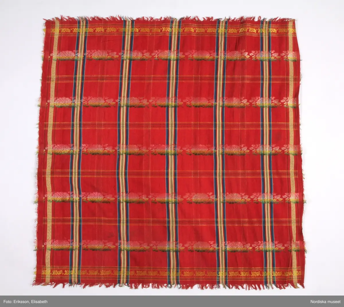 Kvadratiskt halskläde av  halvsiden.  Storrutig  på klarröd botten i satin  med  ränder i schatterande blått, beige, rött och brunt och med jacquardvävda tvärgående blombårder  i rosa, grönt  och gult silke med smala gula  kantbårder. Runtom upprispad kort frans. I ena hörnet en rund svart stämpel med svårläst text: Stockh. Det är Stockholms hallstämpel jfr. likadan t halskläde 44.265 som har en tydligare stämpel  från 1830-talet.
I mycket gott skick.
Troligen en brudschal. Se samma mönster i mörkblått 91.624 från Unnaryd i Småland.
/Berit Eldvik 2012-01-27