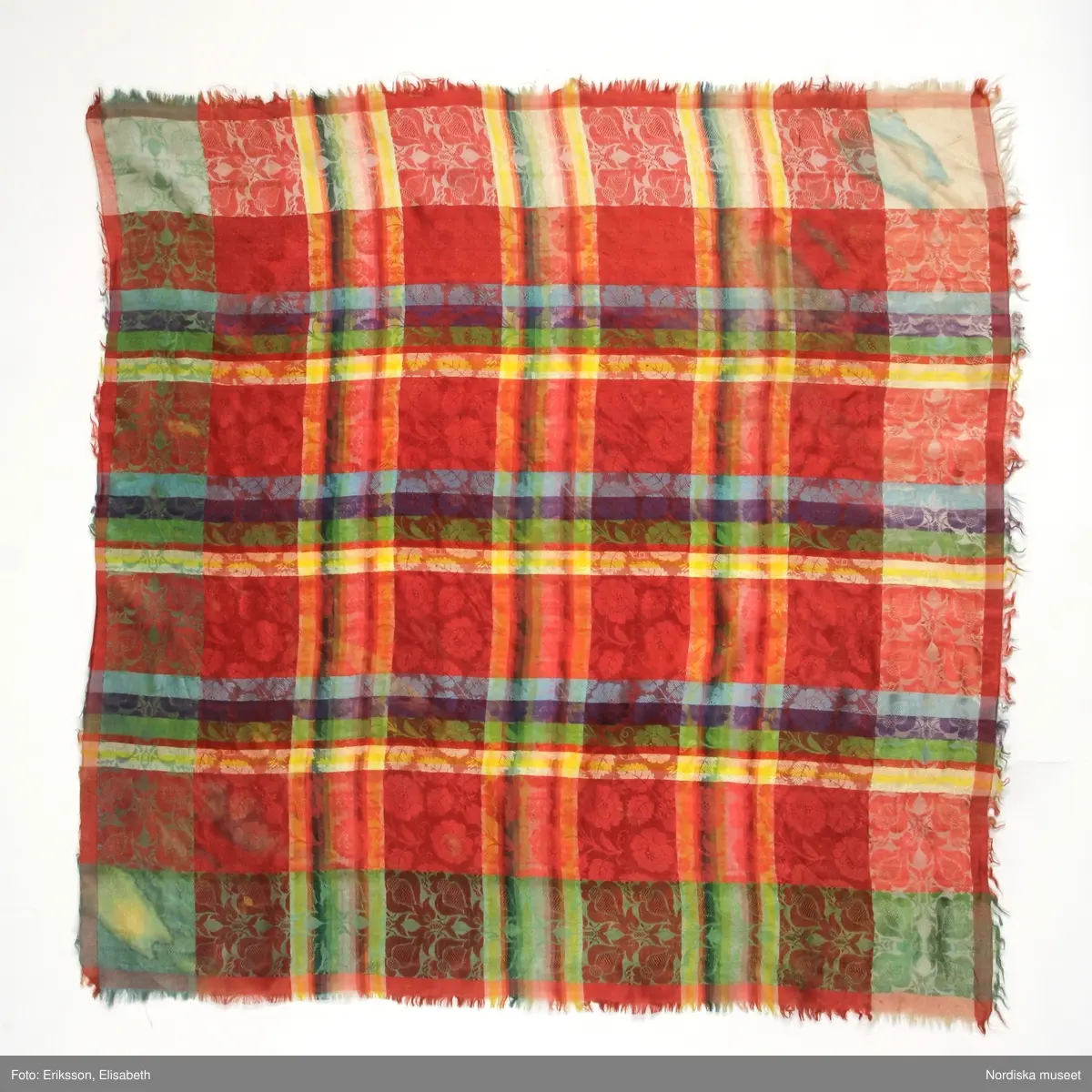 Kvadratiskt halskläde av siden, klarröd botten med rutor i grönt, ljusblått, ljuslila och gult med ett yttäckande invävt mönster, troligen med jacquard, med slingrande rosor. Kantbård med "päronlika" blommor/frukter som på 2 mötande sidor är gröna och på de 2 andra vita. Upprispad kort frans på alla sidor.
I ena hörnet en tydlig svart rund stämpel med texten: Stockh. hallstemp 1839.
Anm. Stor fläck i två av hörnen.
/Berit Eldvik 2011-11-11