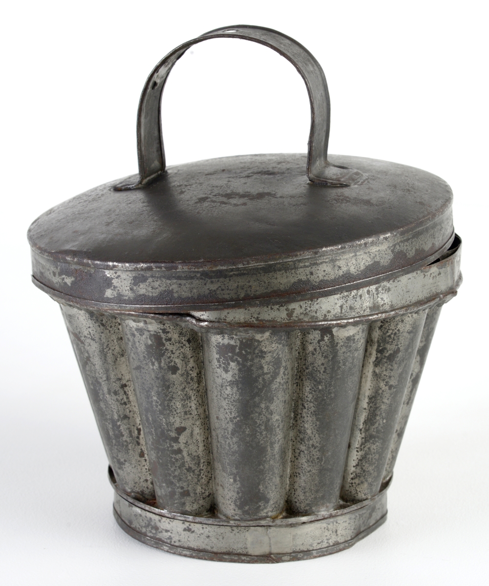 Form med lokk brukt til enten kakebaking eller brødbaking. Formen er rund og har 12 riller på innsiden og 12 tunger rundt kanten. Oppe på formen er det et bånd som holder formen sammen (kun dekor). I bunnen av formen er det en sirkulær fordypning samt en fordypning som forestiller en blomsterstilk og et blomsterblad. Ved hvelving av kaken/brødet vil dette fremstå som et opphøyet mønster på toppen av kaken. Lokket er svart utenpå og grått inni. Lokket har en hank der det er et hull (til oppheng?).