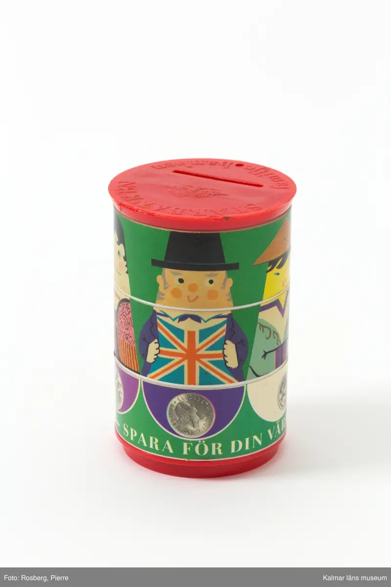 KLM 45519:9 Sparbössa. Rund sparbössa i röd plast. På ovansidan präglat i plasten, Sparbanken, den gamla sparbanksloggan med en ek samt, familjebanken. Här finns också en springa att stoppa pengar i. Sidorna består av tre olika rader som kan vridas i olika lägen. De olika raderna har en klistrad pappersremsa med motiv. Den översta raden har ansikten av människor från olika länder t ex Sverige, England och Kina. Mittdelen utgör människornas torso och nedre delen visar mynt från respektive land. På den nedre delen står, Hela världens sparar – spara för din värld. I botten, präglat i plasten,  MADE IN SWEDEN PAT. PEND. ARTA-PLAST STHLM”.