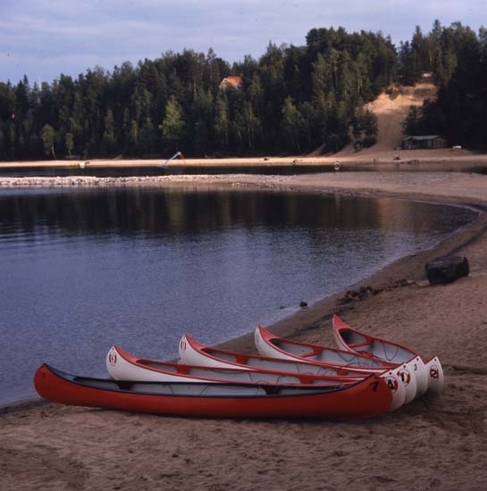 På en sandstrand vid en älv ligger några kanoter uppdragna.