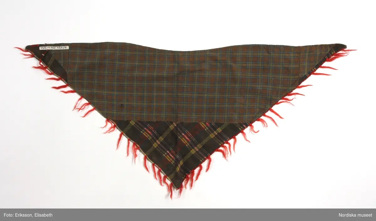 Trekantigt halskläde av troligen jacquardmönstrad bomullssatin.  Grönbrun botten randig i  ljusare grönt, gult och blått med invävd bladslinga i bottenfärgen. Tvärgående mönsterbårder i rött och blått  samt smalare i gult och grönt, vilket ger ett rutigt  tyg. Tvärbårderna i inslagsflotteringar.   Tyget har 2 materialskarvar. I ytterkanterna iknuten gles frans av rött ullgarn.Fodrad till större delen med ett grön/brunrutigt bomullstyg, snibben bak ofodrad.
 Tillverkad av delar från ett kvadratiskt halskläde.
/Berit Eldvik 2012-02-09