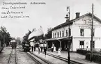 Stationen sedd från spåren med folk utanför. Bredvid ett ensamt lok står tre personer:
1. Stins Cederlund
2. Stionskarl Johnsson
3. Lokförare Samuelsson.