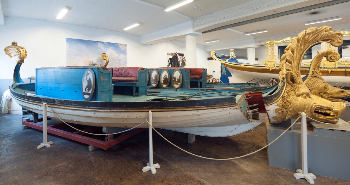In 1783 King Gustaf III travelled to Italy. In Venice he was taken on a tour in a gold-ornamented gallery. The king also watched a splendid spectacle in the form of a regatta on the Canal Grande. A great number of decorated boats and gondolas assembled well before noon. At two o'clock in the afternoon boats with Venetian nobelmen came to pick up the king and his entourage to see the regatta. The boats were decorated with silk, canopies, parasols and flowers. The rowers were dressed in silk and adorned with silver. "The spectacel was vivid, full of splendor and distinctive character and quite dazzling", as Gundmund Jöran Adlerbeth writes in his diary from the journet. King Gustav III was very much impressed.

I order to supreise the king, master ship builder Fredrik Henrik af Chapman had "two pleasure vessels for Haga" built. Haga was a rural ground, north of Stockholm, where Gustav III at the time was creating a royal rank park. The "gondolas" were probably built in 1785 or 1786, and the heads of the boats were in all certainty made by the sculptor Hohan Törnström.

The king was "infinitley pleased", and the sloops came to be used frequently on the Brunnsviken and off Drottningholm. For rowing, boatsmen commandeered from the navy were used. The boar have been called sloops, pleasure vessels or gondolas depending on where they mentioned. The shields were originally 24 in number, with motifs from antiquity and from the twelve labors of Heracles.

After the assassination of Gustav III the sloops were forgotten and rarley used until they were pun into service again for a while during the reign of Karl XV (1859-1872). Since then, they have been stored at various places, e g near the copper tents at Haga, on Skeppsholmen and, since 1966, in the collections of the National Maritime Museum.    