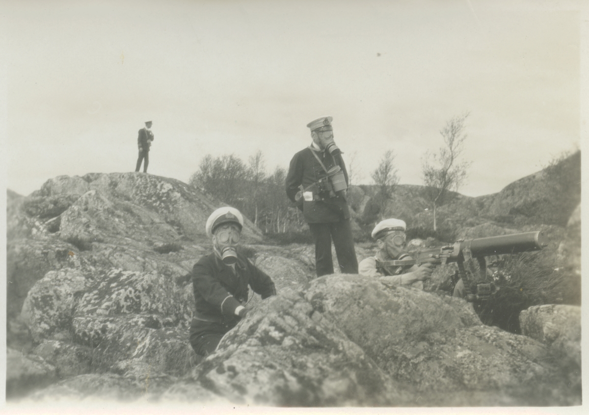 Sjömilitärer i gasmask bemannar kulspruta. Andra bilder i Bergers samling tyder på att fotografiet tagits då officerare och manskap från pansarskeppet SVERIGE 1929 deltog i landstigningsövning på Stabbo i Stockholms södra skärgård. Detta synes bekräftas av att fartygsnamnet kan ses i mössbandet på matrosen vid kulsprutan.