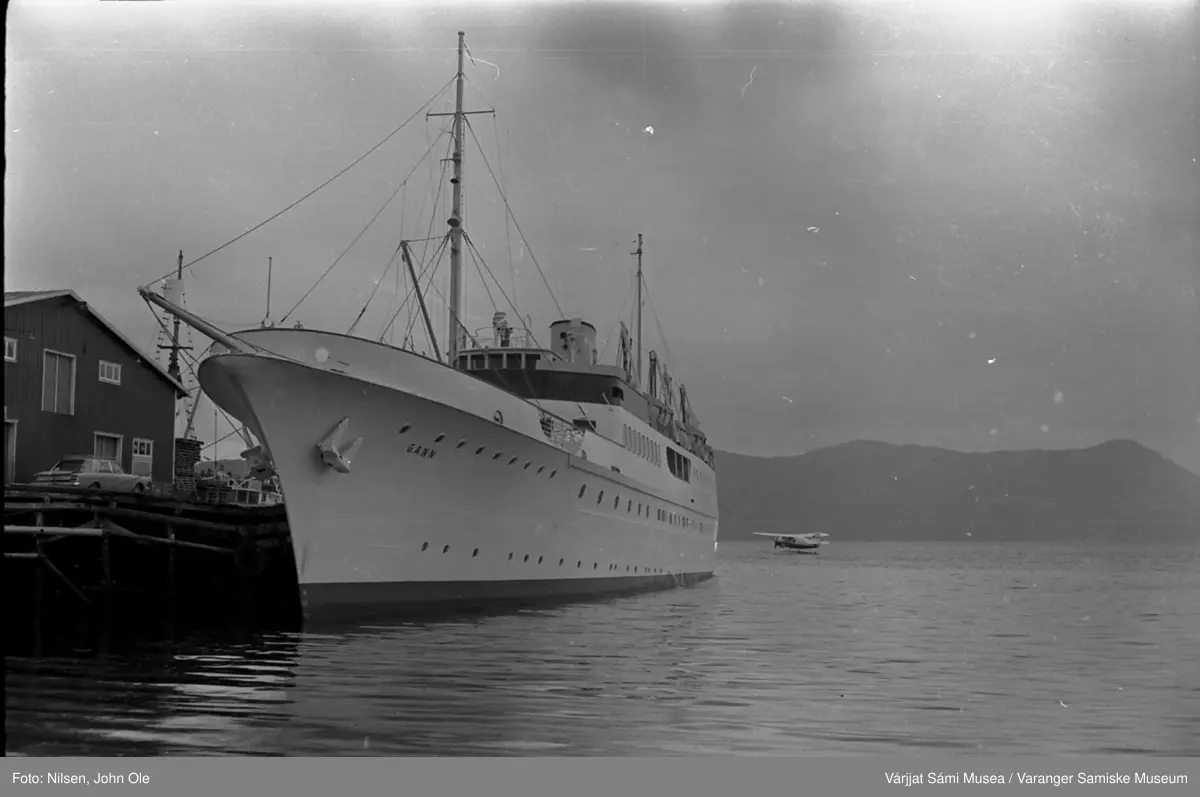 En båt ved navn "Gann" ligger ved kai i Bukta i Alta, et lite sjøfly i bakgrunnen. Alta, Juli 1967