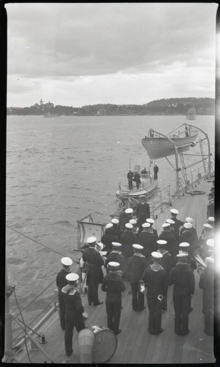 Militärorkester och officerare i paraduniform ombord på pansarskepp (sannolikt SVERIGE) gör sig beredda att ta emot kung Gustaf V, som anländer i motorbarkass. Tid, plats och sammanhang okända. Se även Fo229267C.