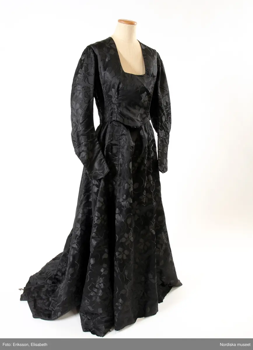 Högtidsklänning med släp, svart jacquardvävt siden, med mönster av blomstergirlanger. Långärmat klänningsliv med fyrkantig urringning. Antydan till fårbogsärm med lätt rynkning vid axelpartierna. Söm mitt på ärmen som kan tyda på att den förlängts från att från början ha varit en 3/4-delsärm. Klockformad kjol, med flera våder, två sömmar på framdelen. Linning ca 2 cm. Liv och kjol är fästade med hakar, men även delvis ihopsydda. Kjollängd fram 105 cm, bak 130 cm. Ärm 60 cm. Midjemått ca 66 cm, ryggmått, livstycke 41 cm. Slitage på utsatta ställen, som nedre kanten och vid linningen. Delvis omsydd. Tryckknappar för knäppning av kjoldelen.
/Karin Dern 2017-05-18
