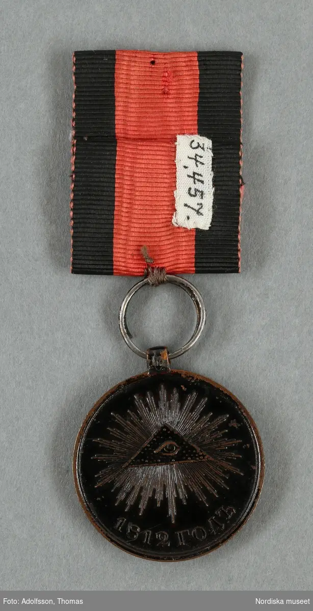 Huvudliggaren:
"Medalj. Se 34.443 [= 34.443-34.485 samlade af friherre Bennet.] 
I tenn, svartmålad. Å ena sidan: Det allseende ögat med strålar; nederst 1812 [och 4 "bokstäver]. Å andra sidan 'HE HAMb, HE MAb ANMEHN TBOEMY [liknande, trol. kyrilliska bokstäver, se huvudliggaren för korrekt avskrift], i fyra rader. Diam. 8 1/2 lin. I rödt, vattradt sidenband med svarta kanter.
Ink. ss. 34.424 [=ink. ss. 34.422  jämte 34.425-34.485]. [34.422=Ink. af byggmästare E. A. Boman i Stockholm 8/1 1883]"