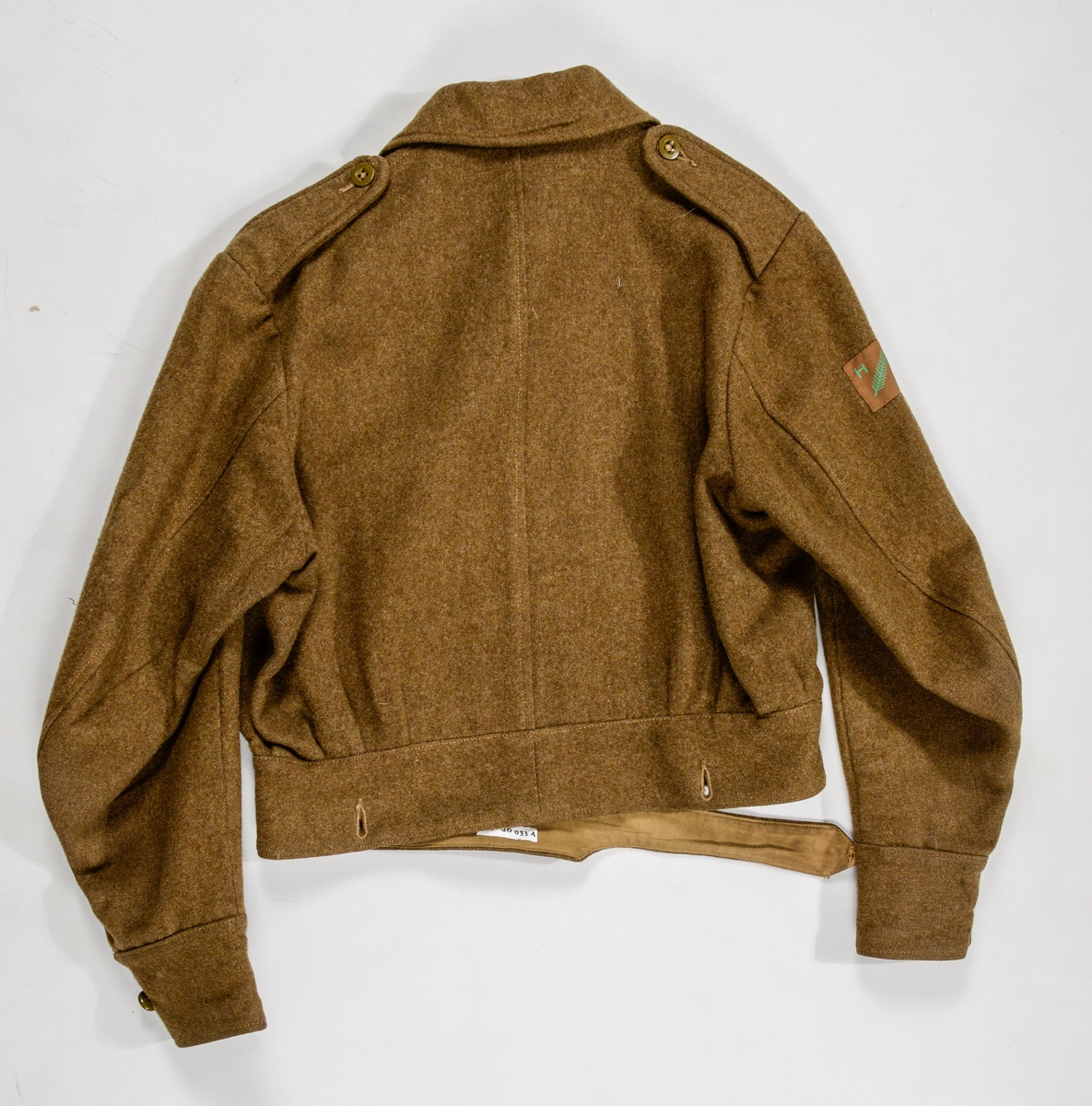 Militæruniform, Jakke (A) i grønt ulltøy. Liggane krage, stolpe i opninga med fem knappehol og fem knappar. Linning nederst med metallspenne. Ei utanpålomme på kvar side med knapp og knappehol. Skulderklaffar. Saum midt bak. Erme med mansjett, knapp og knappehol.
Bukse (B) i same tøy. Linning og gylf med knappar.  To stikklommer, to utanpåsydde lommer på framsida, og ei baklomme.
Armband (C) i gråbrunt med påsydd norsk flagg, to svarte sikkerheitsnåler (var festa til jakka).
Armband (D) i lys brunt med påsydd norsk flagg og to raude stipe (var festa til jakka).
Band (E) Smalt tjukt band (belte) med avrunda metalavslutning i endene. Spenne i eine enden og knapp i den andre.
(C D E ligg i eske i fag III, hylle 2b)