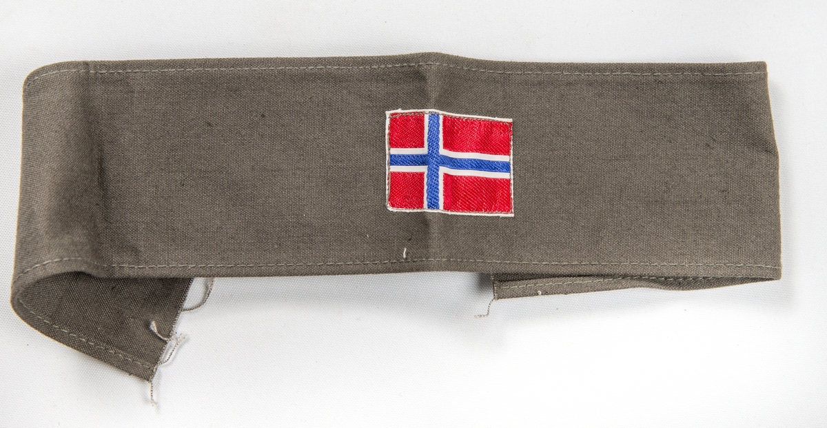 Militæruniform, Jakke (A) i grønt ulltøy. Liggane krage, stolpe i opninga med fem knappehol og fem knappar. Linning nederst med metallspenne. Ei utanpålomme på kvar side med knapp og knappehol. Skulderklaffar. Saum midt bak. Erme med mansjett, knapp og knappehol.
Bukse (B) i same tøy. Linning og gylf med knappar.  To stikklommer, to utanpåsydde lommer på framsida, og ei baklomme.
Armband (C) i gråbrunt med påsydd norsk flagg, to svarte sikkerheitsnåler (var festa til jakka).
Armband (D) i lys brunt med påsydd norsk flagg og to raude stipe (var festa til jakka).
Band (E) Smalt tjukt band (belte) med avrunda metalavslutning i endene. Spenne i eine enden og knapp i den andre.
(C D E ligg i eske i fag III, hylle 2b)