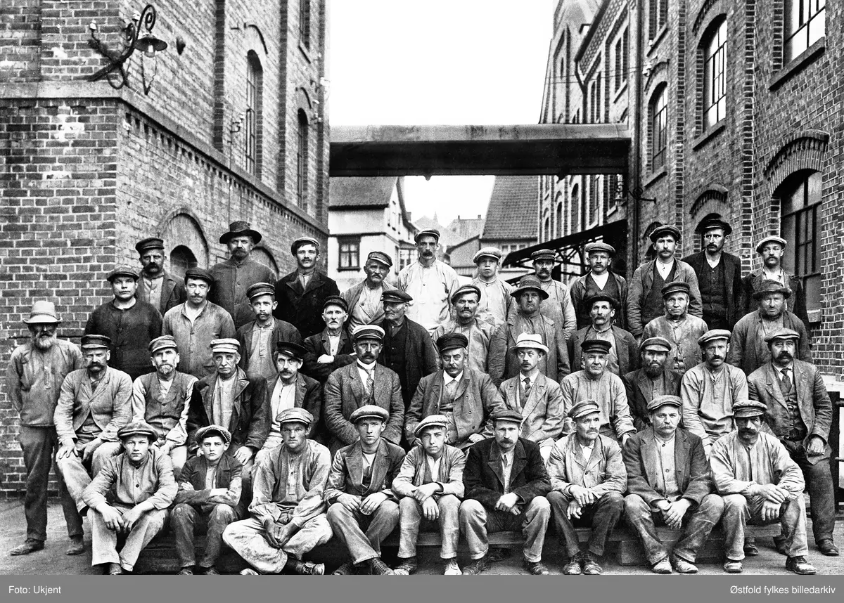 Arbeidsstokken ved Moss Aktiemøller fotografert mellom Central Pakhus og Kloster Mølle i august 1915. 
Siste person på andre rad forfra er Aksel Knudsen (1865-1932). Da dette fotografiet ble tatt var han eldste funksjonær i Moss Aksjemøller, og formann ved firmaets nye og moderne maismølle.