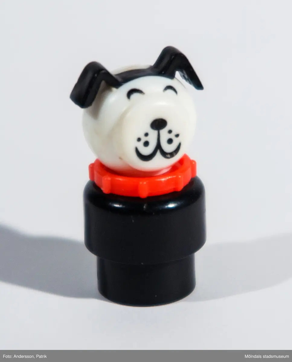Rund hundfigur av plast tillhörande Fisher-Pricegarage från 1970-talet. "Kroppen" består av två delar varav den nedersta är smalare för att passa ihop med tillhörande leksaksbil. Ihålig underifrån och längst in är "huvudet" fastskruvat. Mellan kropp och huvud sitter ett hundhalsband. "Huvudet" består av en något tillplattad plastkula med markerade hundöron samt målade ögon, morrhår, nos och mun. Inköpt 2001 på Second Hand av givaren, vars barn har lekt med det.