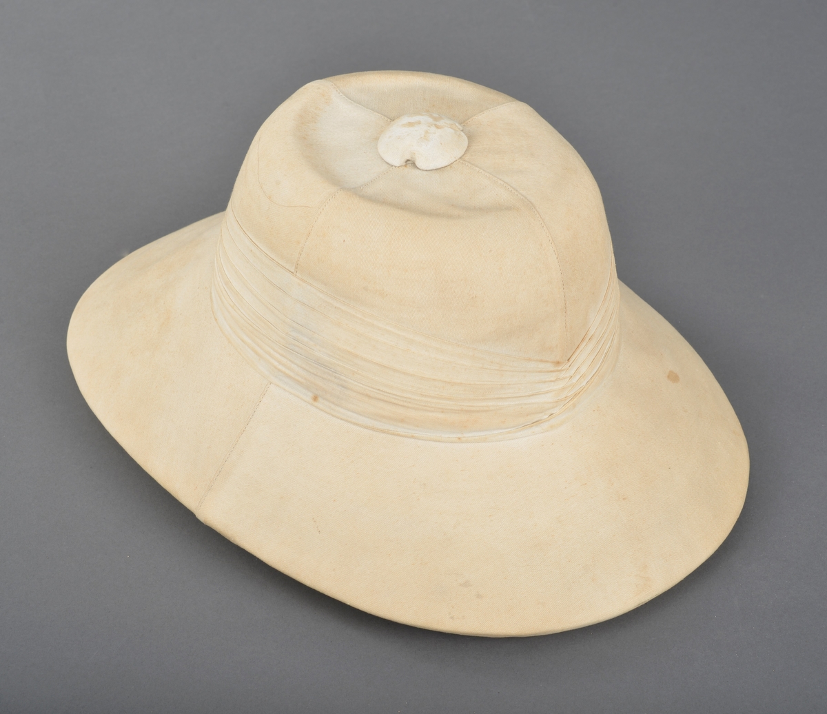 Hatt trekt med stoff. Pullen er sydd sammen av fire deler. Trekt knopp på toppen. Rundt pullen er hatten dekorert med flere lag med stoff. Pullen og bremmen er av lett og tykt materiale. Pullen er foret og i toppen er det trykt fabrikkmerke. Bremmen er foret med grønt stoff. Skinnkant er festet i nedere kant av pullen.