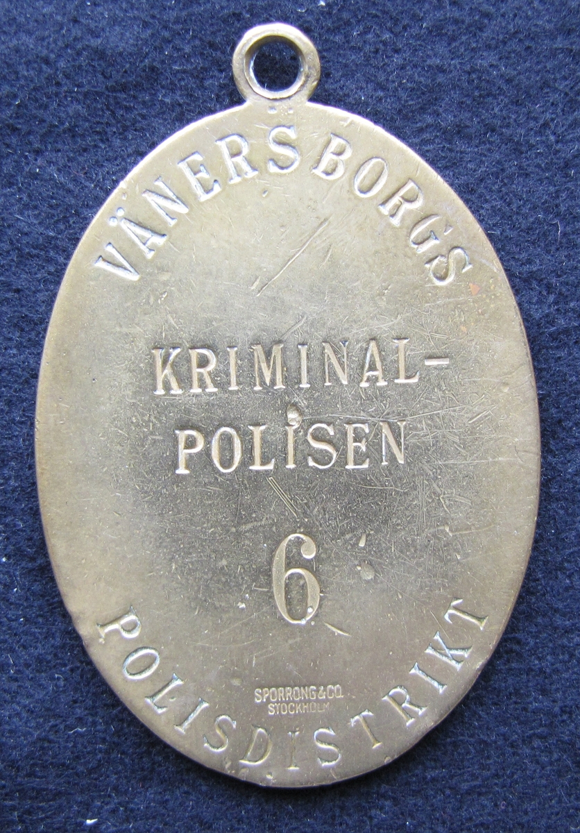 Polisbricka, oval med en ögla i toppen. 

På framsidan: Lilla riksvapnet, dvs en krönt sköld med tre kronor. Bakom skölden eklöv och korslagda fasciner. Under emblemet står det POLIS.

På frånsidan: VÄNERSBORGS POLISDISTRIKT står den i det övre och undre kanten. Mitt på står KRIMINALPOLISEN och siffran 6. Även tillverkaren Sporrong & CO, Stockholm finns ingraverat.