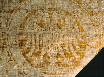 Mässhaken är vävd i s.k. Regensburg-brokad. Dubbelt varpsystem med lin och silke, botteninslag av silke samt mönsterinslag av tarmguld spunnet runt en lintråd. (Taquete alt. samitum)
Tarmguldet är idag bortnött.

Mönstret i form av dubbelörnar inom cirklar har stark anknytning till italiensk-bysantinska mönster. 

Enligt A. Geijer tillverkad i Spanien eller Tyskland.

Litteratur: Medeltida vävnader och broderier i Sverige.
av A. Branting och A. Lindblom. del II, plansch 217, text sid 101 ff.
"Ur Textilkonstens historia", Agnes Geijer. 1972. (sid 162, pl 34 a.)