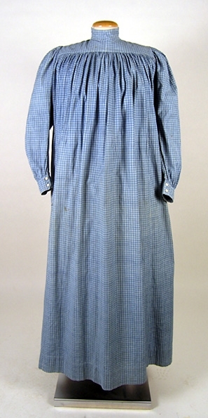 Klänning (bakförkläde) av blå-vit rutigt bomullstyg. Klänningen är rynkad runt om det släta oket. Oket är fodrat med ett vitrandigt bomullstyg. Manschetterna har sprund och knäpps med 2 trädda knappar. Framtill på höger sida finns ett hål för en ficka. I midjan finns en dragsko som knyts framtill på magen på insidan. Klänningen knäpps baktill med 7 hakar och sydda hyskor. Ett 11 cm brett skärp i samma tyg knytes runt midjan. Fläckar framtill samt ett hål.