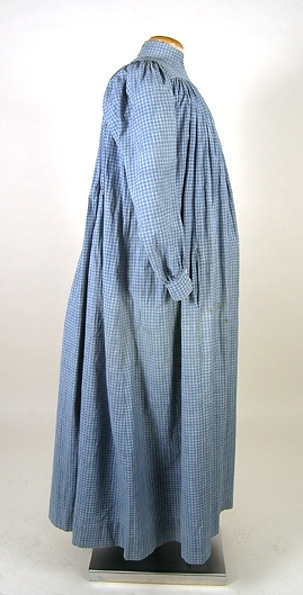 Klänning (bakförkläde) av blå-vit rutigt bomullstyg. Klänningen är rynkad runt om det släta oket. Oket är fodrat med ett vitrandigt bomullstyg. Manschetterna har sprund och knäpps med 2 trädda knappar. Framtill på höger sida finns ett hål för en ficka. I midjan finns en dragsko som knyts framtill på magen på insidan. Klänningen knäpps baktill med 7 hakar och sydda hyskor. Ett 11 cm brett skärp i samma tyg knytes runt midjan. Fläckar framtill samt ett hål.