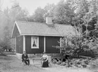 Liten stuga, herre och dam sittande framför. 

Fotograf: Ellen Kock? Axel Lundbäck m.fl.
Gullspångssamlingen.
Bilder från Gullspång med omnejd.