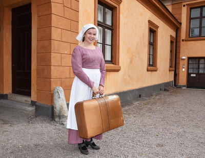 En jente utkledd som tjenestepike står ute i portrommet med en koffert i hånda. 