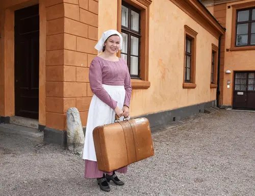 En jente utkledd som tjenestepike står ute i portrommet med en koffert i hånda. 