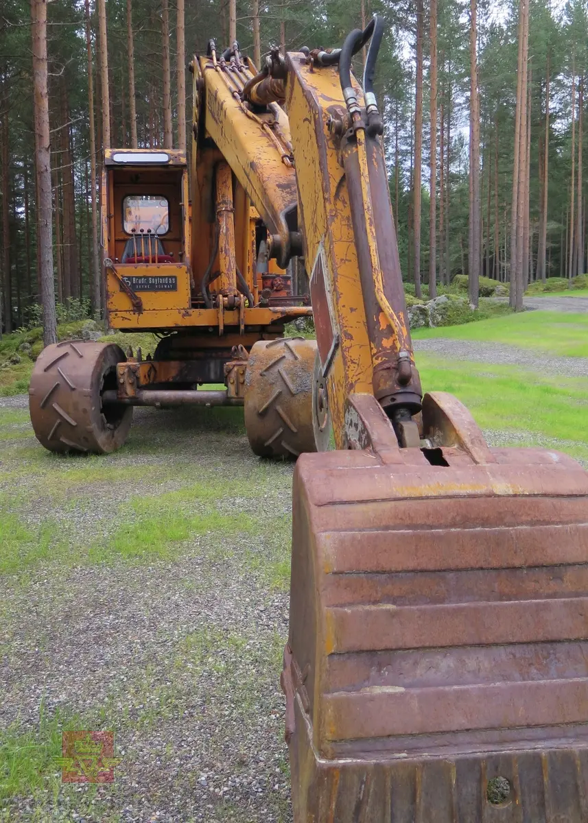 Gravemaskin, hjulgående. 2 jernhjul foran og 2 gummihjul bak. Maskinen er oransje med svart sete. Motoren er en 4-sylindret dieseldrevet type fra Perkins. Gravemaskinen var ikke selvgående, men forflyttet seg med gravearmen under arbeid. Ved transport mellom arbeidsstedene hektet en skuffen i lasteplanet og løftet opp "forhjulene" slik at den ble transportert som en henger.