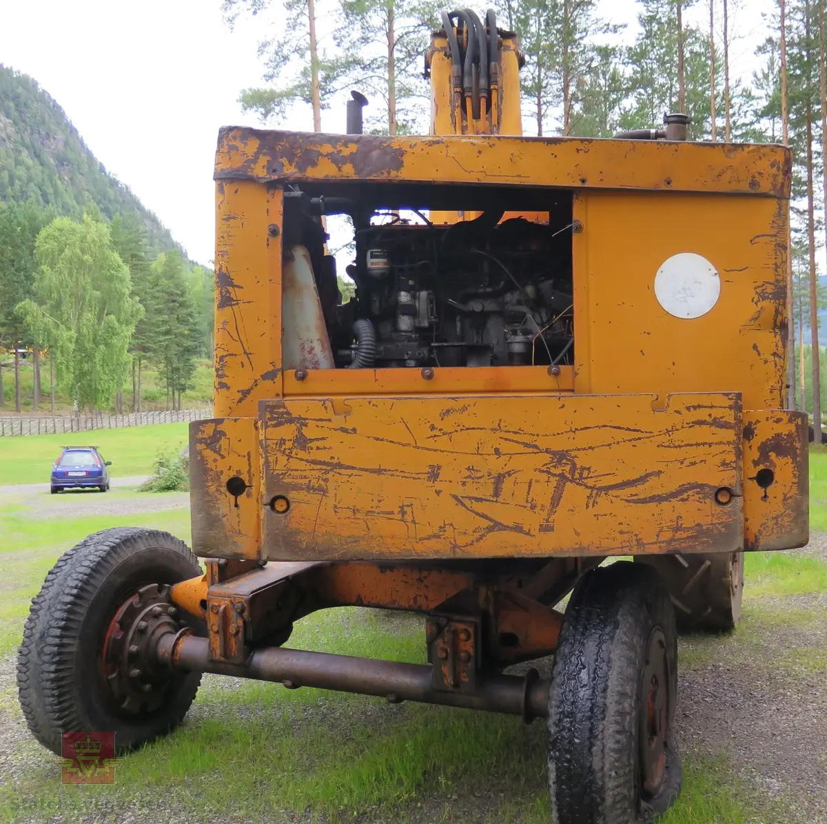 Gravemaskin, hjulgående. 2 jernhjul foran og 2 gummihjul bak. Maskinen er oransje med svart sete. Motoren er en 4-sylindret dieseldrevet type fra Perkins. Gravemaskinen var ikke selvgående, men forflyttet seg med gravearmen under arbeid. Ved transport mellom arbeidsstedene hektet en skuffen i lasteplanet og løftet opp "forhjulene" slik at den ble transportert som en henger.
