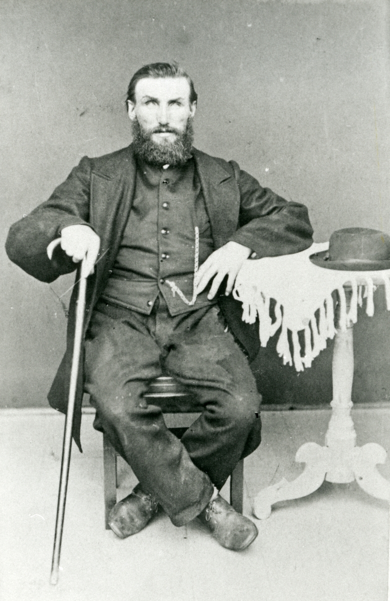 Mann sittende m/stav ved siden av bord. Hatt oppå bordet. Portrett av Anders Engemoen f. Moldberge (1834)