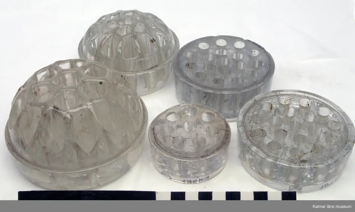 KLM 41862 Blomhållare, 7 st, glas. Rund med flera hål, olika storlek och form. 2 st med kupad ovansida, oregelbundna hål, den största med diameter 11 cm, höjd 7 cm, den mindre med diameter 8 cm, höjd 6 cm. 5 st med platt ovansida, runda hål, den största med diameter 9 cm , höjd 3,5 cm, den minsta med diameter 6 cm, höjd 2,5 cm. Använd placerad i botten på en vas eller skål, i hålen sticks blomstjälkar ner.