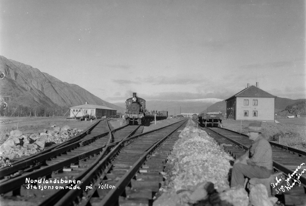 Under anleggsperioden på jernbanestasjonen.
Til venstre Reisegodsekspedisjonen, til høyre stasjonen.
Det første lokomotivet som kom til Mosjøen står på stasjonen. Bakpå ser vi mange mann i arbeid.
I forgrunnen mann som sitter på stein.