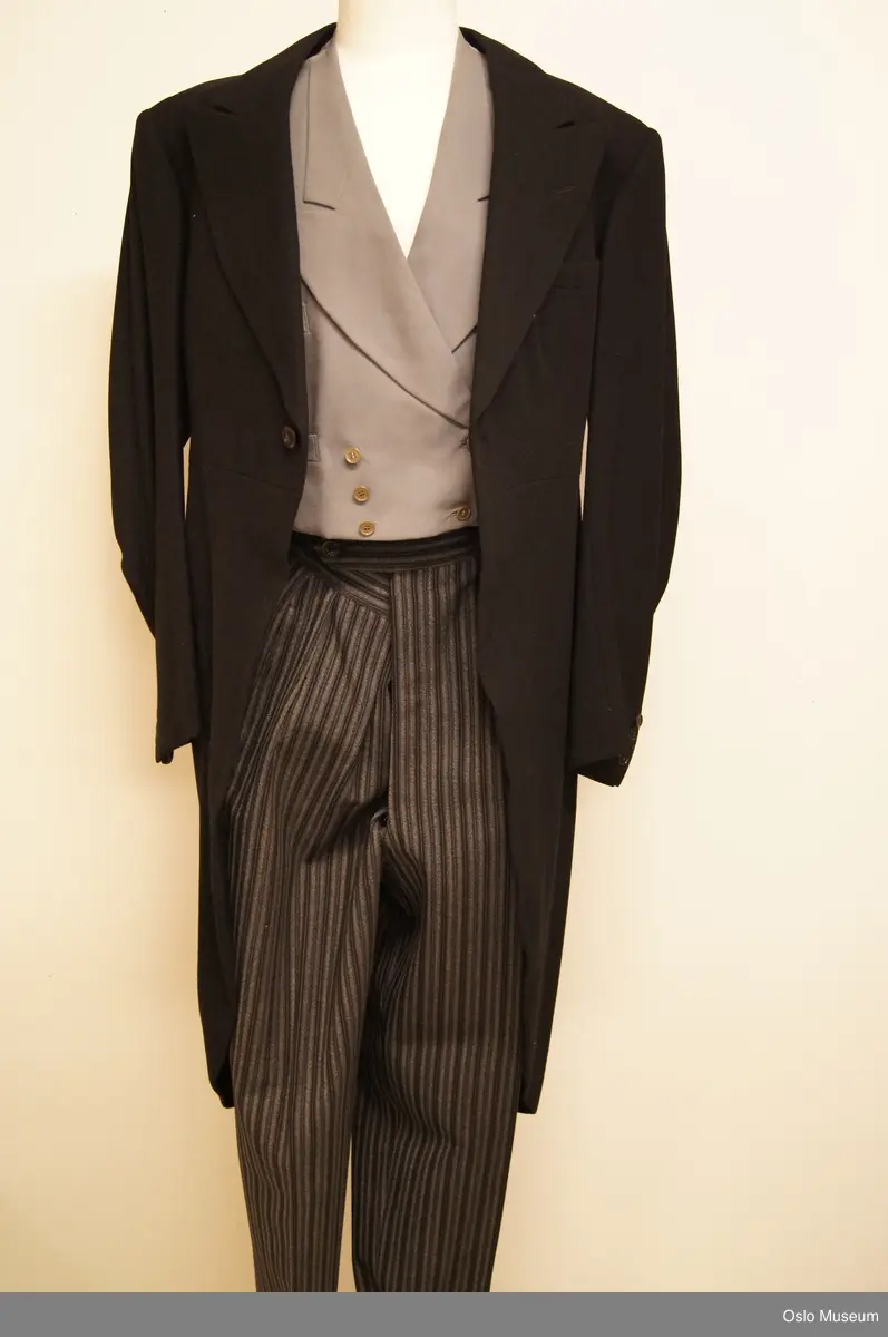 A: Sjakett av sort fiskebensvevet ullstoff. Kraven rundet bak, går over i rettvinklet avslutning foran. Brede, spisse slag. Venstre side med stikklomme. Fra livsømmen strekt utskrådde flak som går over i parti med rundet nedkant. Splitt midt bak, knapp i livet. Lange ermer, nederst med splitt og fire knapper, fóret med sort silke, ermene med hvit silke, skulderputer. B: Buksen av sort og hvitstripet ull, rynket til linningen ,lukning med knapper. Linningen fóret med elfenbensfarget silke.To stikklommer og to baklommer av sort bomullstoff.  C: Vest i grått ullstoff. Rundet nakke med krave. Lange,spisse slag, dobbeltknappet med tre knapper, innsnitt. Fire stikklommer. Ryggdel av grå silke, fór av elfenbensfarget silke To innerknapper. D: Vest av stoff som jakken. Spiss ringning, knapping med seks knapper. Innsnitt og lommer og fór som på C.