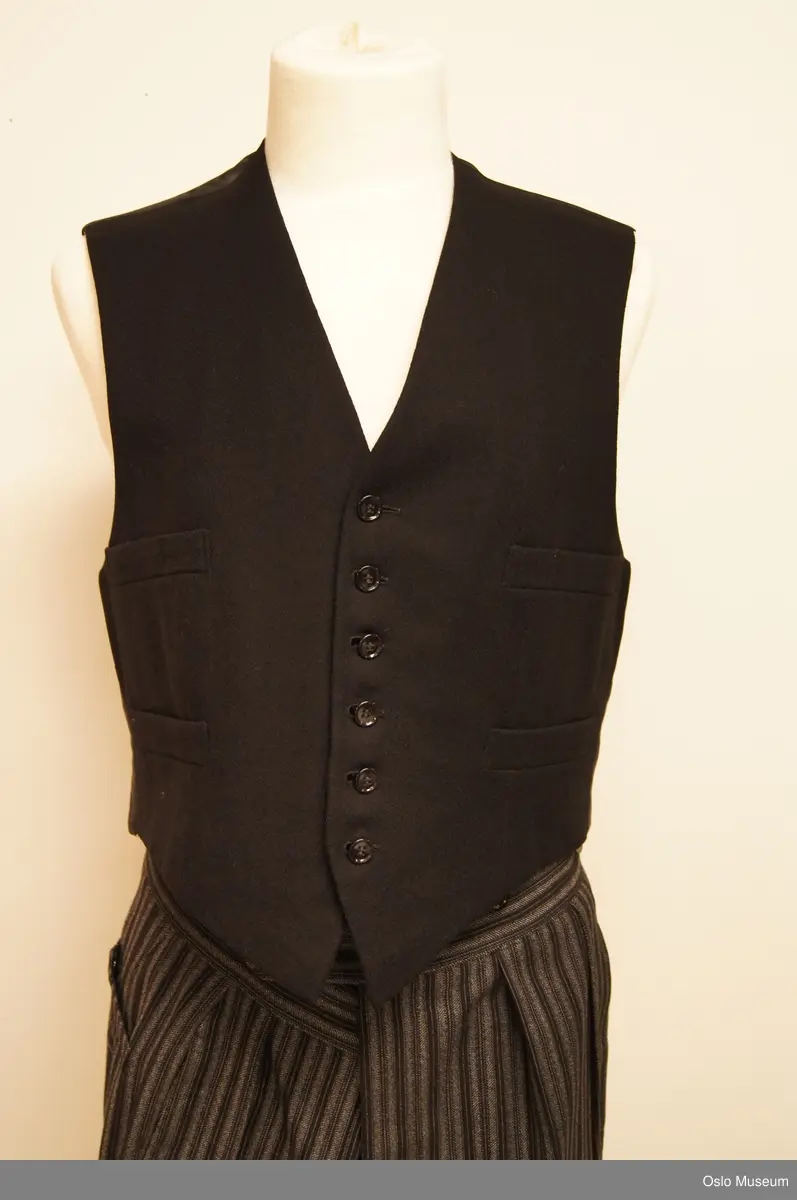 A: Sjakett av sort fiskebensvevet ullstoff. Kraven rundet bak, går over i rettvinklet avslutning foran. Brede, spisse slag. Venstre side med stikklomme. Fra livsømmen strekt utskrådde flak som går over i parti med rundet nedkant. Splitt midt bak, knapp i livet. Lange ermer, nederst med splitt og fire knapper, fóret med sort silke, ermene med hvit silke, skulderputer. B: Buksen av sort og hvitstripet ull, rynket til linningen ,lukning med knapper. Linningen fóret med elfenbensfarget silke.To stikklommer og to baklommer av sort bomullstoff.  C: Vest i grått ullstoff. Rundet nakke med krave. Lange,spisse slag, dobbeltknappet med tre knapper, innsnitt. Fire stikklommer. Ryggdel av grå silke, fór av elfenbensfarget silke To innerknapper. D: Vest av stoff som jakken. Spiss ringning, knapping med seks knapper. Innsnitt og lommer og fór som på C.