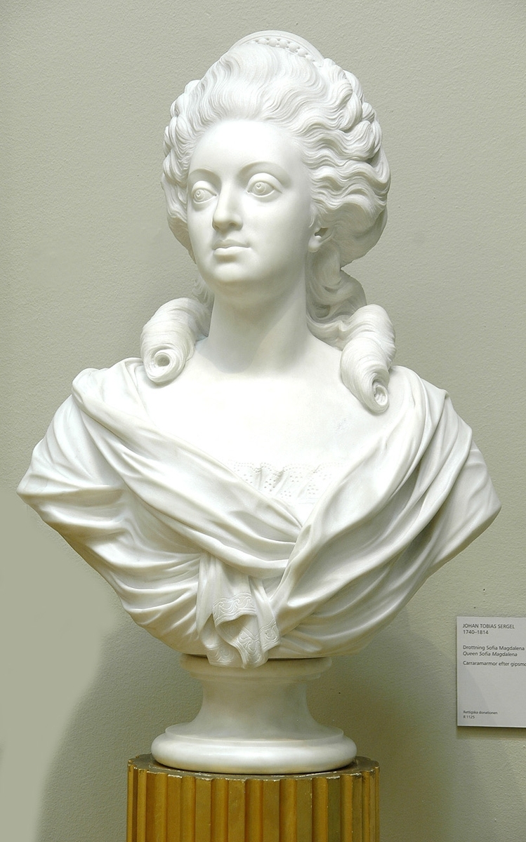 Marmorbyst, föreställande Sveriges drottning Sofia Magdalena. "Copia fatta da M. Bacci, Florens", dvs kopia av Bacci efter J T Sergels gipsoriginal, år 1783. 
Står på hög gustavians piedestal, med vertikala hålkäl och lagerkrans nertill kring foten. Piedestalen står på lågt fyrkantigt podium.