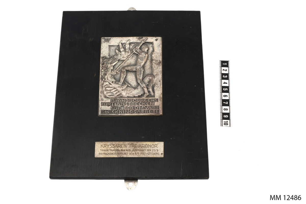 Av silver, på platta av svartpolerad ek. I relief, bild av naken yngling i strid med en eldsprutande drake, därunder följande text: Brandbolagens förtjänsttecken för berömvärt släckningsarbete. På bricka på plattan, texten: Kryssaren Tre Kronor Timber trading den 10/8, Järntorget den 25/8 och magasinkvarteret den 4/9 1947 Göteborg.